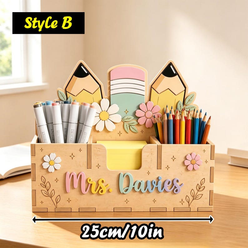 Op de afbeelding: Een houten bureau-organizer van 25 cm of 10 inch breed, met de tekst "Mrs. Davies" in pastelkleurige letters. Het achterpaneel toont twee gele potloden en een gum, versierd met witte en roze bloemen en bladeren als decoratieve elementen.