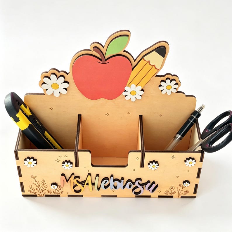Op de afbeelding: Een houten bureau-organizer met een uitgesneden rode appel en geel potlood aan de bovenkant. De voorzijde is versierd met witte madeliefjes en kleine sterretjes. De naam Ms. Alebiosu staat in cursief schrift. Het bevat een stanleymes en een schaar.
