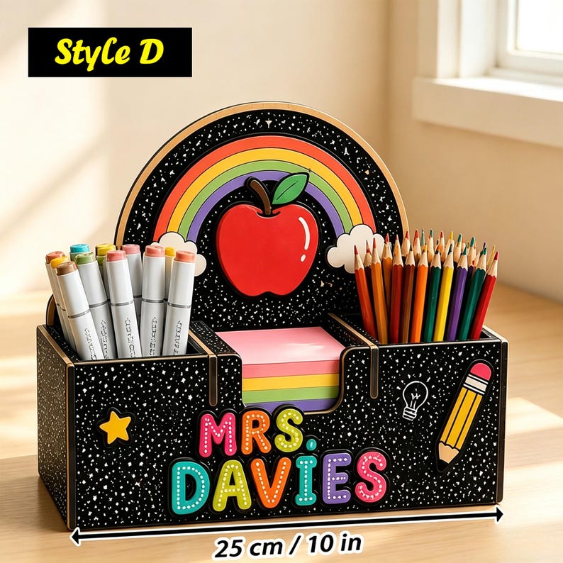 Op de afbeelding: Een zwarte bureau-organizer met spikkels, voorzien van een regenboog, rode appel, gele ster, gloeilamp en potloodillustratie. De tekst luidt 'MRS. DAVIES'. Het bevat stiften en kleurpotloden en is 25 cm breed.