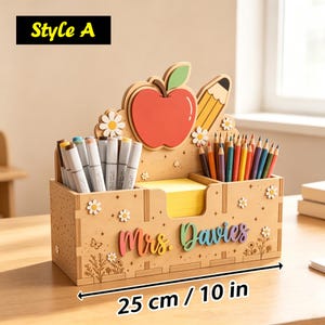 Op de afbeelding: Een houten bureau-organizer van 25 cm breed, voorzien van een rode appel, potlood, madeliefjes en de tekst 'Mrs. Davies'. Het bevat vakken met stiften en kleurpotloden, plus een centraal geel blokje met zelfklevende notitieblaadjes.