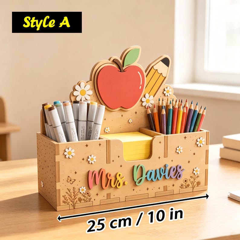 Op de afbeelding: Een houten bureau-organizer van 25 cm breed, voorzien van een rode appel, potlood, madeliefjes en de tekst 'Mrs. Davies'. Het bevat vakken met stiften en kleurpotloden, plus een centraal geel blokje met zelfklevende notitieblaadjes.
