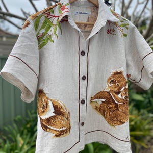 May include: A beige short-sleeved button-down shirt featuring a brown koala print on the front, green eucalyptus leaf patterns near the collar, and dark brown piping along the edges. The shirt has a label reading 'notretree' and size 'L'.