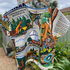 May include: A short-sleeved top made from a vintage-style tea towel featuring a colourful print of Coonabarabran, Australia. The design includes a Tyrannosaurus, an observatory, a courthouse, and printed text including a poem and beer branding.