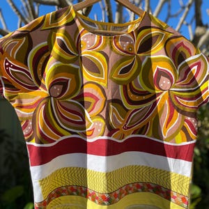May include: A short-sleeved top featuring a vibrant floral pattern in shades of yellow, brown, and pink. The lower section displays a horizontal red stripe, a white band, and a yellow border with a geometric pattern and small colourful accents.