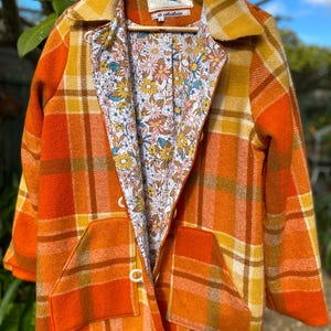 May include: A long-sleeved jacket featuring an orange, yellow, and white plaid pattern on the exterior. The interior is lined with a white fabric displaying a multi-coloured floral print. The jacket includes two front pockets and white buttons.