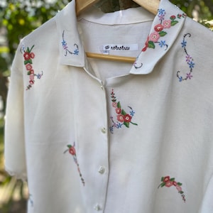 May include: A cream-coloured short-sleeve button-down shirt featuring delicate floral embroidery in shades of pink, red, and blue. The shirt has a classic collar and a white interior label with the text "notnotnew" and the size "M" printed on it.