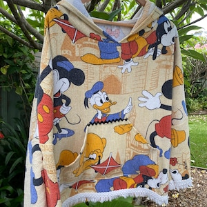 May include: A cream-coloured hooded sweatshirt with a white fringe trim. The sweatshirt features a colourful all-over print of Mickey Mouse, Donald Duck, and Goofy.
