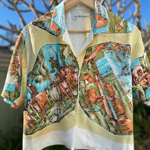 May include: A short-sleeved button-down shirt in pale yellow featuring a vibrant, multi-coloured print of Australian landscapes, cattle, and windmills. The collar displays map outlines of Darwin and Tasmania. The shirt has a white hem and clear buttons.