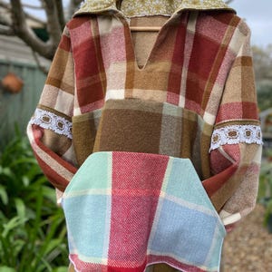 May include: A long-sleeved pullover shirt featuring a multi-coloured plaid pattern in shades of brown, red, and cream. The garment includes a large front pocket with a light blue and red plaid design, white lace trim on the sleeves, and a collar lined with a green floral fabric.