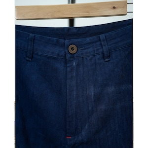 May include: Close-up of dark blue denim pants. The pants have a button closure, belt loops, and a small red stitch detail. The fabric appears to be a medium weight denim.