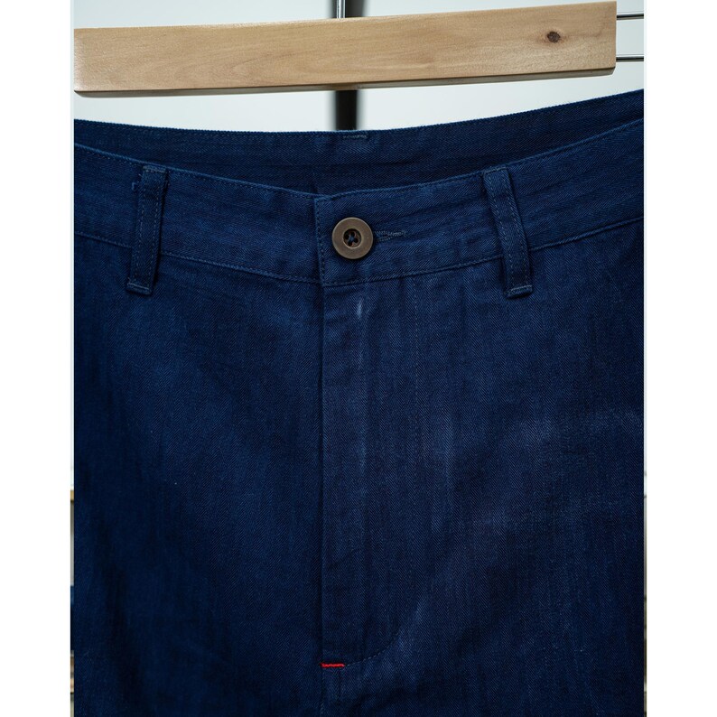 May include: Close-up of dark blue denim pants. The pants have a button closure, belt loops, and a small red stitch detail. The fabric appears to be a medium weight denim.