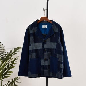 May include: A navy blue jacket with a patchwork design in shades of blue and grey. The jacket features a collared neckline, three buttons, and two front pockets. The sleeves are a solid navy blue.