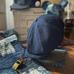 Puede incluir: Gorra de estilo newsboy azul marino con un patrón de punto texturizado. La gorra se exhibe sobre un soporte de madera, con un silbato de latón vintage colgando de un cordón de cuero. Otros sombreros de mezclilla están en el fondo.
