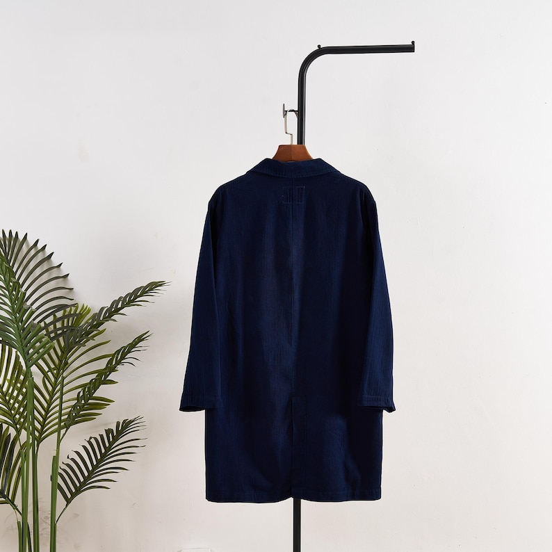 May include: A navy blue long coat with a collared neckline and long sleeves hangs on a black metal clothing rack. The coat is made of a solid, dark blue fabric and appears to be a lightweight garment.