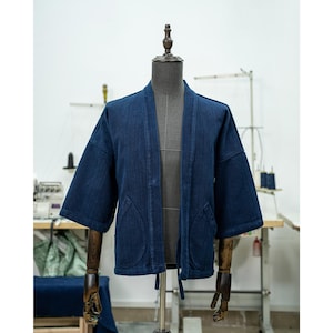May include: A dark blue denim jacket in a traditional Japanese style, with a wide collar, long sleeves, and a tie closure at the waist. The jacket has two large pockets on the front.