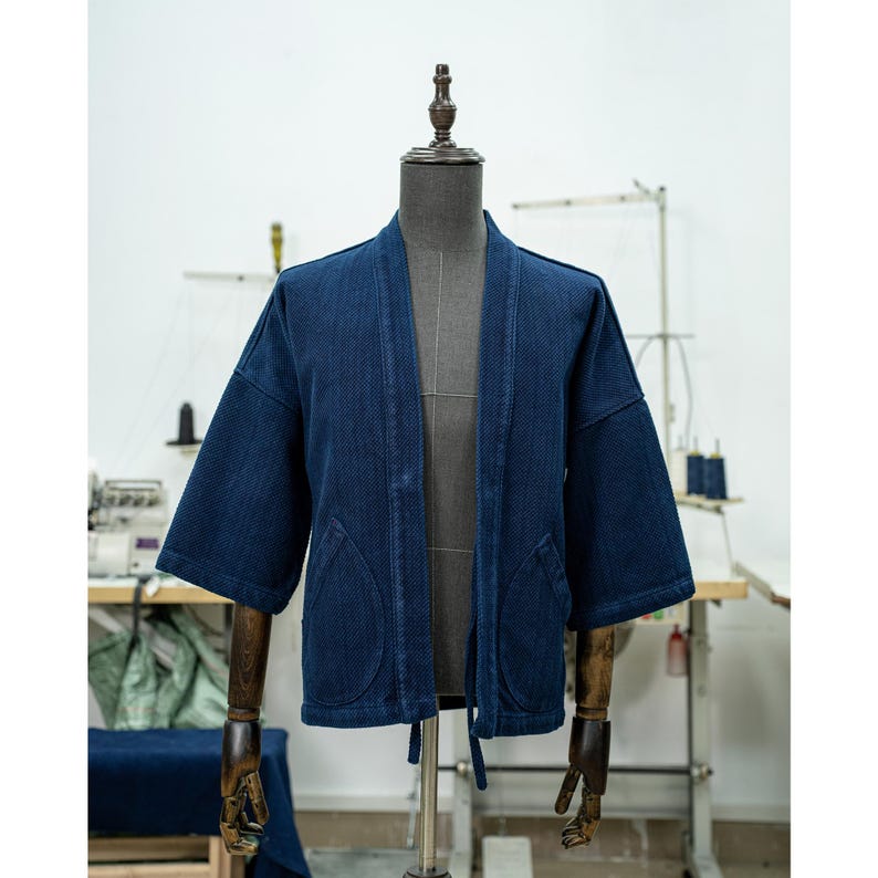 May include: A dark blue denim jacket with a traditional Japanese style, featuring a wide collar, long sleeves, and a tie closure at the waist. The jacket has two large pockets on the front.