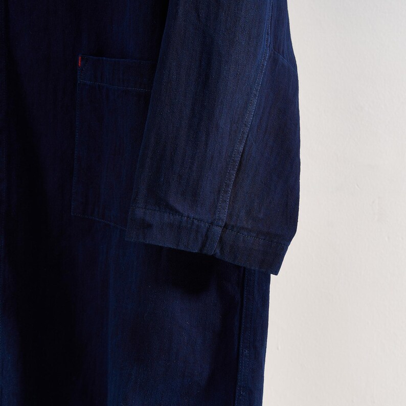 May include: Close-up of a dark blue denim jacket with a single pocket and a red accent. The jacket has long sleeves and is made of a textured fabric. The garment is likely part of a clothing collection.