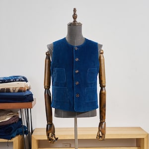Puede incluir: Un chaleco de pana azul marino con cuatro bolsillos delanteros y botones de madera. El chaleco se exhibe en un maniquí. El chaleco es de color azul oscuro liso. El maniquí tiene brazos de madera.