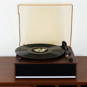 May include: A dark wood-grain record player with a black front speaker panel and a white base trim. It features a black vinyl record on the platter with a gold logo, a black tonearm, two black control knobs, and a translucent amber dust cover.