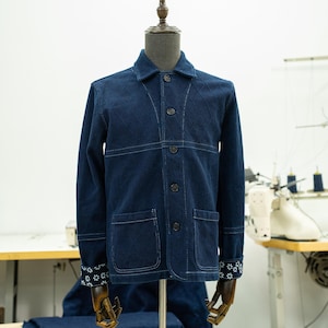 Puede incluir: Una chaqueta vaquera azul oscuro con un patrón acolchado de rombos y costuras blancas. La chaqueta tiene un cierre de botones en la parte delantera, dos bolsillos delanteros y bordados florales blancos en los puños.