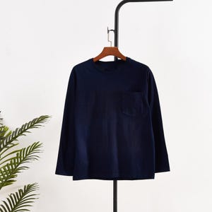 Könnte beinhalten: Ein marineblaues Langarmshirt mit Brusttasche. Das Shirt hängt an einem Holzbügel. Das Shirt ist aus einem weichen, bequemen Material gefertigt.