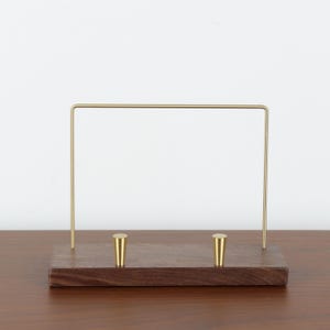 May include: A rectangular dark brown wooden base featuring two small, upright, conical brass pegs. A thin, U-shaped brass wire frame is attached to the back of the wooden base, extending upward to create a display stand for items.