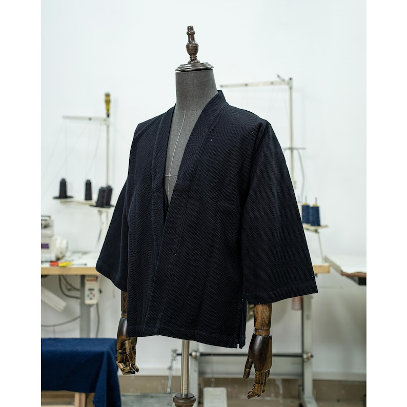 May include: A black cotton kimono jacket with a long, open front and three-quarter sleeves. The jacket is on a mannequin with wooden arms.