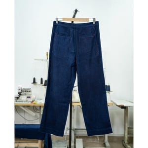 May include: Dark blue denim pants hanging on a wooden hanger. The pants have a wide leg design and red stitching details. The background shows a sewing machine and a work table, suggesting a garment workshop.