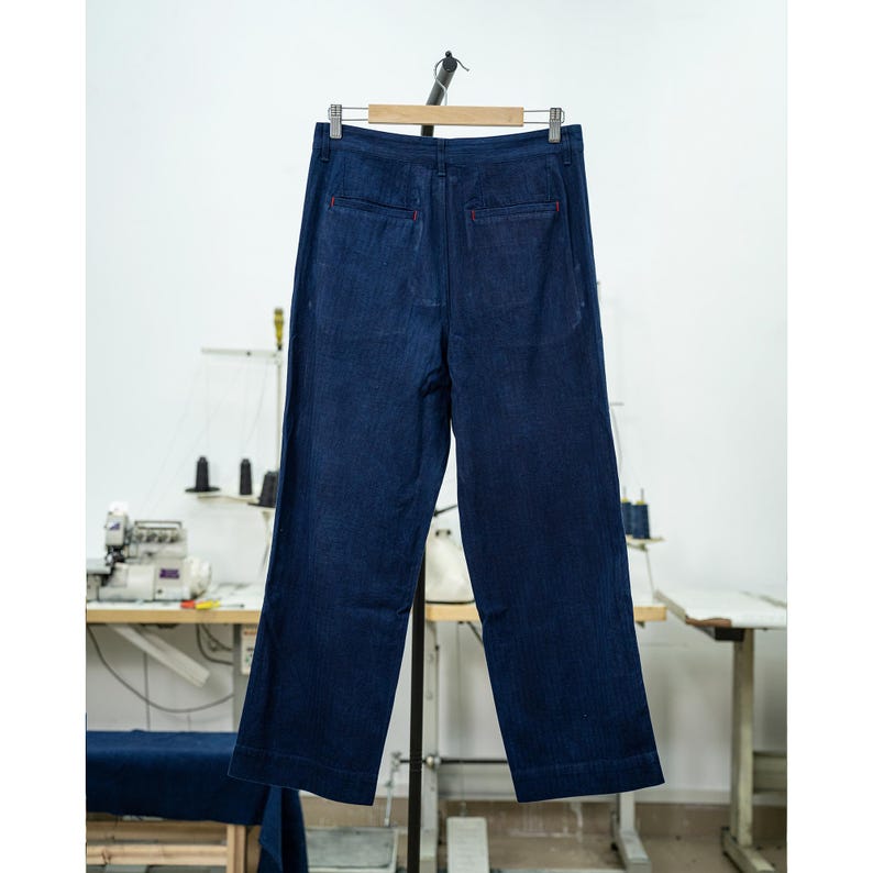 May include: Dark blue denim pants hanging on a wooden hanger. The pants have a wide leg design and red stitching details. The background shows a sewing machine and a work table, suggesting a garment workshop.