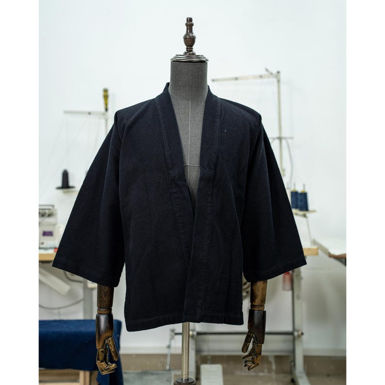 May include: A black, open-front jacket with long sleeves and a textured fabric. The jacket is displayed on a mannequin.