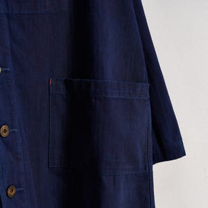May include: A navy blue button-down shirt with a single chest pocket. The shirt has a relaxed fit and features visible stitching details. The buttons are a light brown color. A small red accent is visible on the pocket.