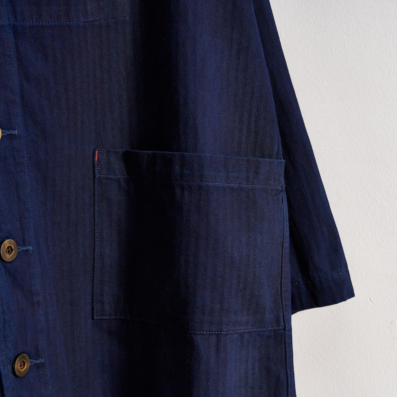 May include: A navy blue button-down shirt with a single chest pocket. The shirt has a relaxed fit and features visible stitching details. The buttons are a light brown color. A small red accent is visible on the pocket.
