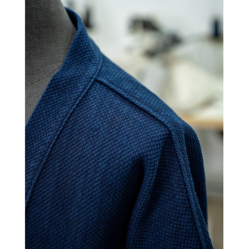 May include: Close-up of the shoulder seam of a dark blue indigo cotton jacket. The fabric has a textured weave.