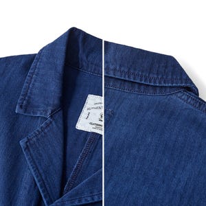 May include: Close-up of a dark blue denim jacket. The jacket features a notched lapel, visible stitching, and a white tag with the words "ORIGINAL AUTHENTIC CLOTHING". The fabric has a textured appearance.