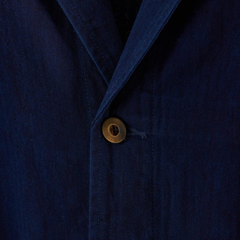 May include: Close-up of a navy blue blazer with a single, round, brass-colored button. The fabric has a subtle texture, and the button is centered on the jacket's front closure. The blazer's lapel is visible.