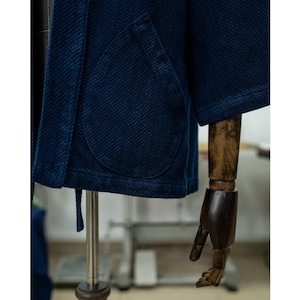May include: A dark blue indigo cotton jacket with a single patch pocket on the left side. The jacket has a traditional Japanese style with a tie closure.