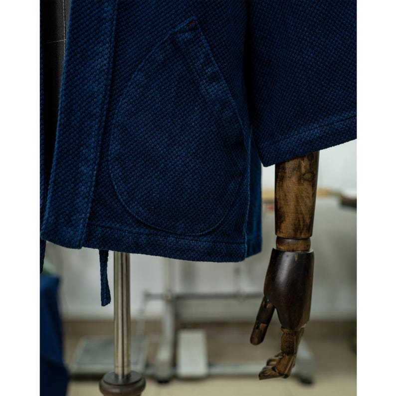 May include: A dark blue indigo cotton jacket with a single patch pocket on the left side. The jacket has a traditional Japanese style with a tie closure.