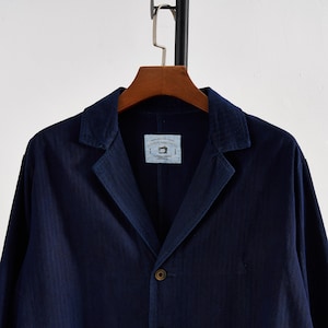 May include: A navy blue blazer with a notched lapel and a single-button closure. The jacket hangs on a wooden hanger. A small tag is visible on the inside of the jacket with the text "AUTHENTIC HANDCRAFTED".