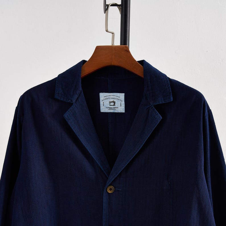 May include: A navy blue blazer with a notched lapel and a single-button closure. The jacket hangs on a wooden hanger. A small tag is visible on the inside of the jacket with the text "AUTHENTIC HANDCRAFTED".