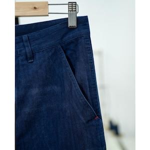 May include: Close-up of dark blue denim pants hanging on a wooden hanger. The pants have a classic design with a front pocket and a red stitch detail. The fabric appears to be a medium weight denim.