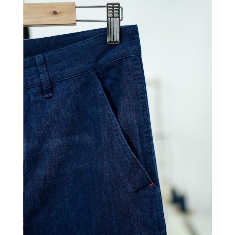 May include: Close-up of dark blue denim pants hanging on a wooden hanger. The pants have a classic design with a front pocket and a red stitch detail. The fabric appears to be a medium weight denim.