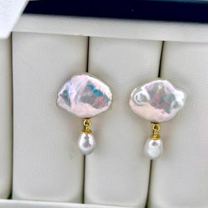 May include: A pair of elegant earrings featuring large, irregular-shaped, iridescent pearls with a gold-colored metal accent. Each earring has a smaller, teardrop-shaped pearl dangling below. The earrings are displayed in a white jewelry box.