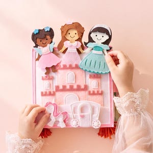 May include: A felt princess playset featuring a pink castle, a white carriage, and a unicorn. Three felt princess dolls in pink, blue, and brown are included. The playset is held by a person wearing a white lace sleeve.