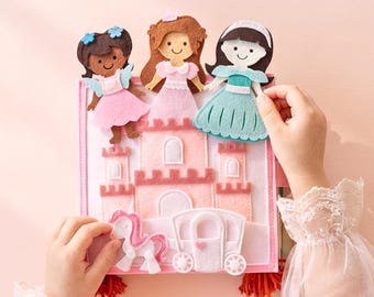 Könnte beinhalten: Ein quadratisches Filz-Aktivitätsbuch mit einem rosa Schloss, einem weißen Einhorn und einer weißen Kutsche. Drei Filzpuppen mit unterschiedlichen Haarfarben und Kleidern sind oben befestigt. Das Buch besteht aus geschichteten Filzteilen in Rosa, Weiß, Braun und Türkis, mit roten Quasten an den unteren Ecken.