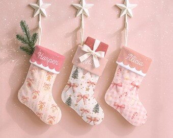 May include: Three personalized Christmas stockings with star-shaped hangers. Each stocking has a different pattern and color scheme, including pinks, whites, and gold. The stockings are decorated with bows, trees, and gingerbread men, with names like "Harper", "Yyla", and "Alicia".