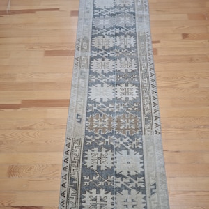 May include: A long, narrow rug with a beige background and a geometric pattern in shades of brown, blue, and white. The rug has a fringe on both ends.