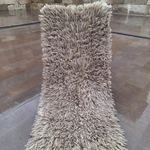 Peut inclure: Un tapis rectangulaire et touffu dans les tons beige et marron. Le tapis a un poil épais et texturé, créant une apparence douce et accueillante. Le tapis est posé sur une surface en pierre mouillée, avec un mur en pierre en arrière-plan.