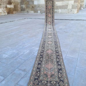 May include: A long, narrow rug with an intricate pattern of floral and geometric designs. The rug features a central field of peach and cream hues, bordered by a dark brown and green pattern. The rug is displayed outdoors, showcasing its length and detail.