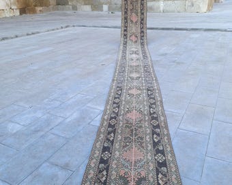 May include: A long, narrow rug with an intricate pattern of floral and geometric designs. The rug features a central field of peach and cream hues, bordered by a dark brown and green pattern. The rug is displayed outdoors, showcasing its length and detail.