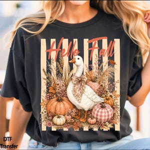 May include: A dark gray t-shirt featuring a graphic print of a white goose wearing a patterned neck bow, surrounded by pumpkins, wheat, and dried foliage. The design includes vertical beige stripes and the orange text Hello Fall.